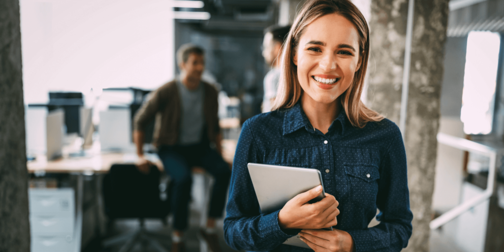 Smiling professional holding a tablet in a bright open-plan office, with coworkers collaborating in the background—representing a productive and modern office environment.