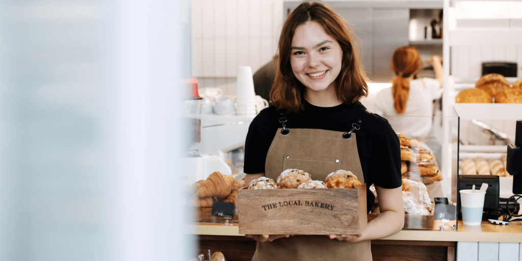 Smiling female bakery worker holding a wooden crate of assorted pastries inside a modern bakery counter area.