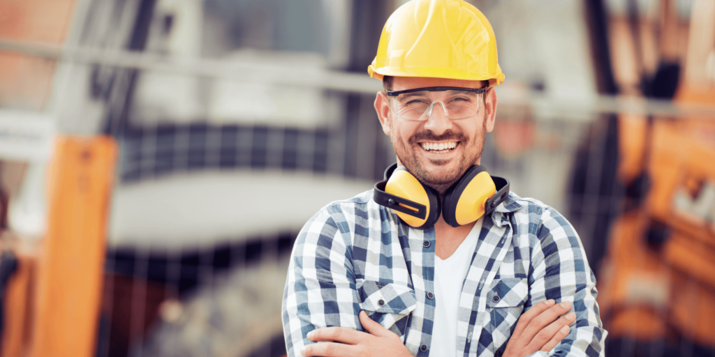 Confident builder in a hard hat and safety goggles smiling on a construction site, wearing a check shirt and protective ear defenders.