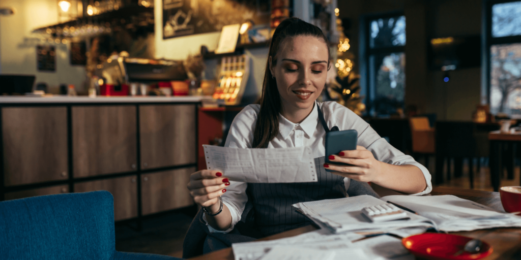 A young woman in a café or small restaurant reviews receipts and paperwork while checking her phone, suggesting she's managing business finances or doing bookkeeping.
