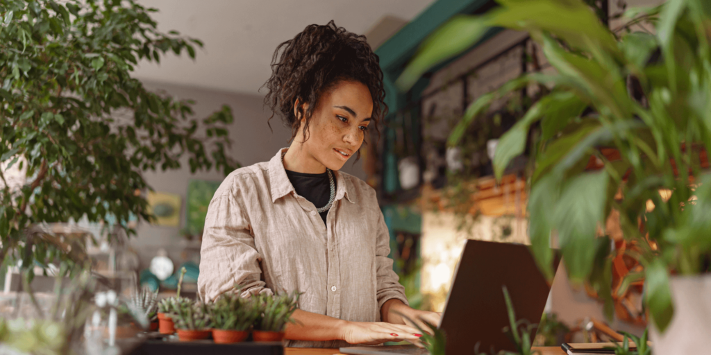 Female florist using a laptop in a plant-filled florist shop, with potted plants and greenery surrounding her workspace.