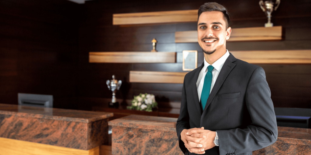 Smiling hotel receptionist or manager in a dark suit standing confidently at a modern hotel reception desk with wooden décor and trophy displays.