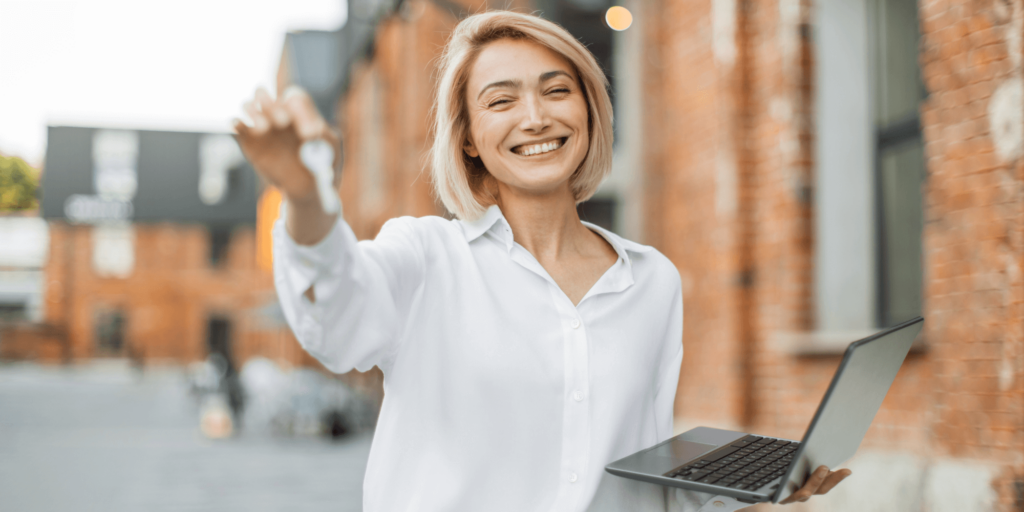 A smiling woman in a white blouse holding a set of keys and a laptop, standing outside a red-brick residential building, symbolising a confident residential landlord or property manager.