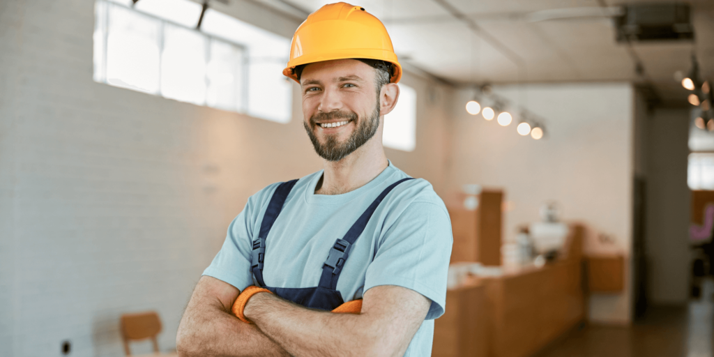 Smiling tradesman wearing a yellow hard hat and overalls standing confidently inside a modern worksite or completed commercial interior.