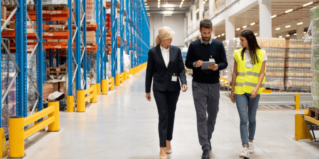 Three people walking in a warehouse, discussing while looking at a tablet, one wearing a safety vest.
