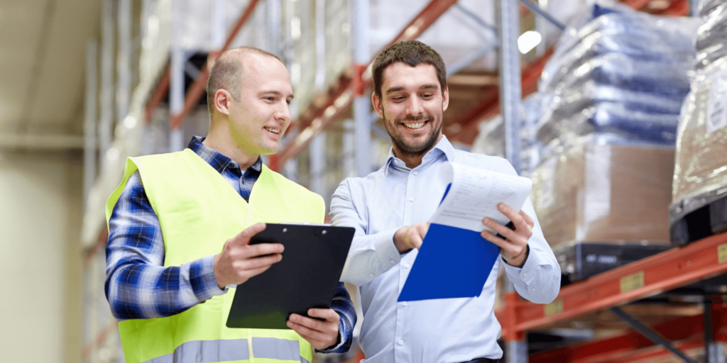 Two warehouse workers smiling while reviewing documents and clipboards in a storage facility.